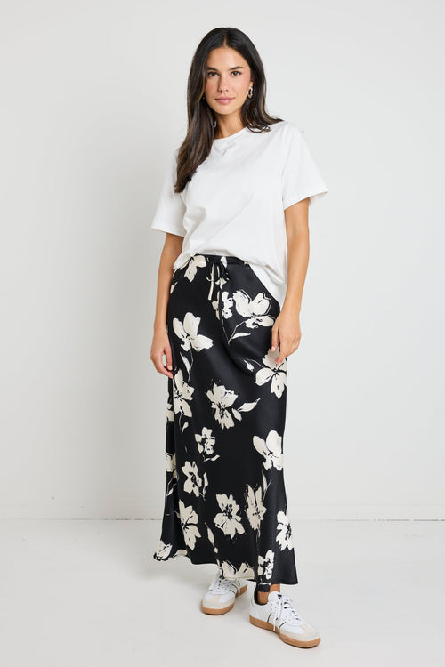 Woman wearing a black floral satin maxi skirt with a tie waist, paired with a white tee for an effortlessly chic and elegant look.