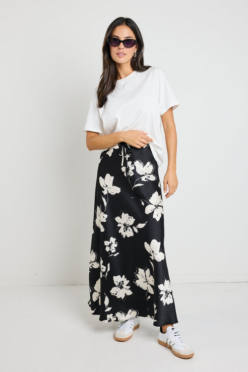 Woman wearing a black floral satin maxi skirt with a tie waist, paired with a white tee for an effortlessly chic and elegant look.