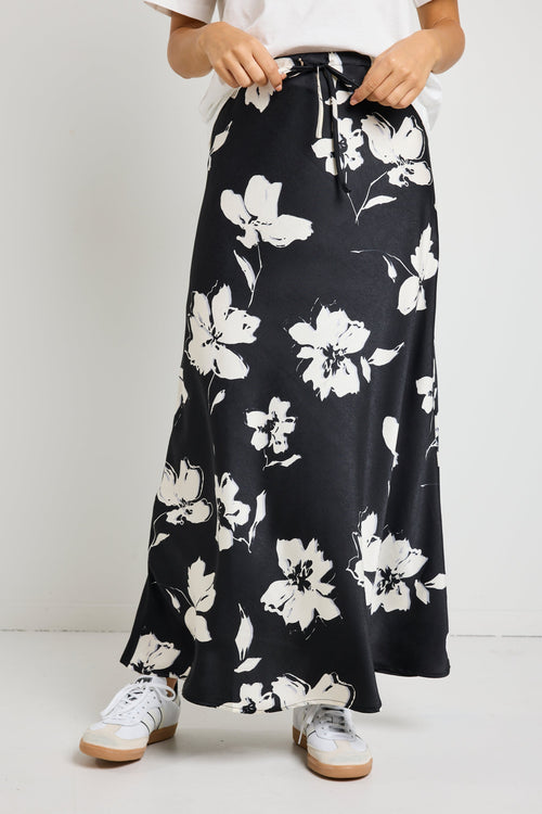 Woman wearing a black floral satin maxi skirt with a tie waist, paired with a white tee for an effortlessly chic and elegant look.