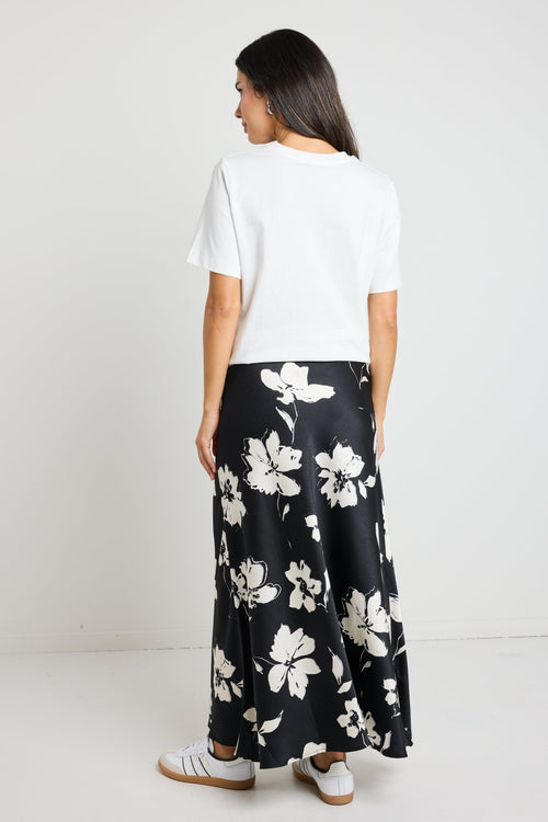 Woman wearing a black floral satin maxi skirt with a tie waist, paired with a white tee for an effortlessly chic and elegant look.