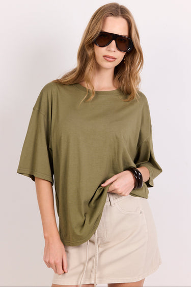 A woman wearing an oversized olive green short-sleeve t-shirt and a beige mini skirt, styled with dark sunglasses and bracelets, against a plain light gray background.