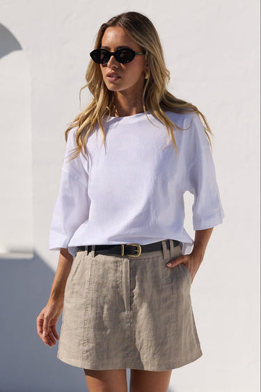 A woman wearing a white off-the-shoulder top, a beige linen mini skirt, a black belt, and black sandals, standing in a sunny, minimalist white-walled courtyard.