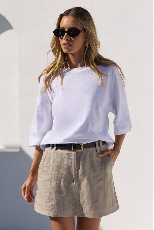 A woman wearing a white off-the-shoulder top, a beige linen mini skirt, a black belt, and black sandals, standing in a sunny, minimalist white-walled courtyard.