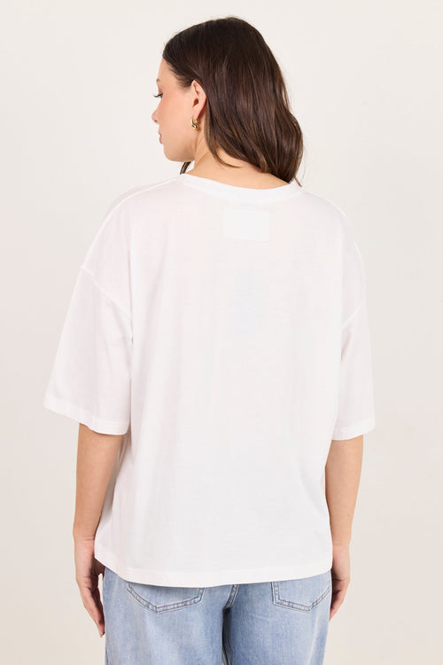 A woman viewed from the back wearing an oversized white short-sleeve T-shirt and light blue jeans, standing against a plain off-white studio background.