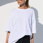 A woman modeling a white oversized short-sleeve top and black wide-leg lace pants, holding a red handbag, set against a minimalist white architectural background with strong sunlight and shadows.