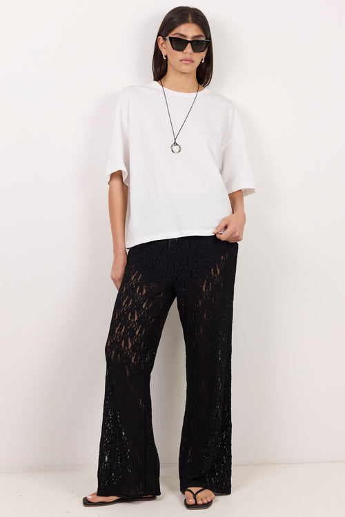 A model stands against a plain white wall wearing a white oversized t-shirt, black wide-leg lace-up pants, black sandals, and sunglasses.
