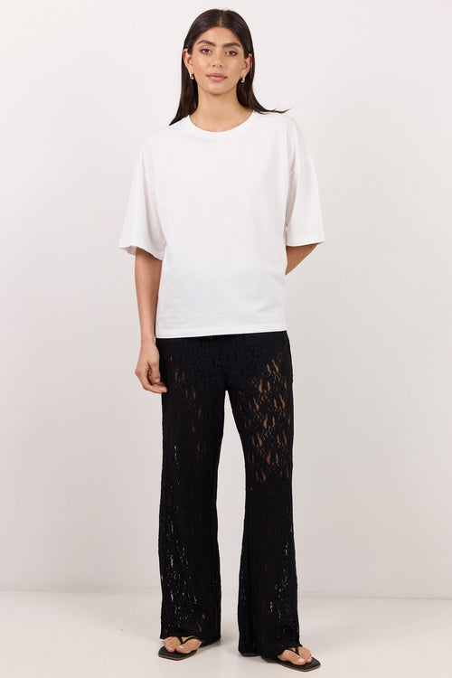 A woman stands against a plain off-white studio background wearing a loose white short-sleeve T-shirt and black wide-leg lace-up pants with a sheer floral lace overlay, paired with dark sandals.