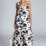 Woman wears an ivory maxi dress with a bold black floral print, wide straps, V-neckline, back tie detail, and a flowing skirt with side splits. The look is fresh, feminine, and relaxed.