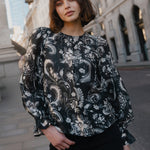 Woman wearing a black floral blouse standing in an urban setting with classical architecture.