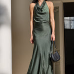 A woman wearing a dark green satin halter-neck maxi dress stands on stone steps outside a building with white columns, holding a dark blue-gray handbag.