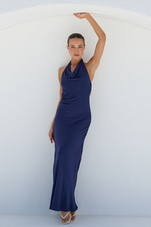 A woman modeling a sleeveless navy blue maxi dress with a draped neckline, posing in front of a white arched wall.
