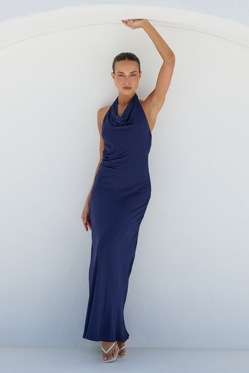 A woman modeling a sleeveless navy blue maxi dress with a draped cowl neckline, posing inside a white arched structure.