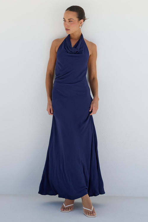 A woman modeling a sleeveless navy blue halter-neck maxi dress with a draped cowl neckline, standing against a minimalist white arched studio backdrop.