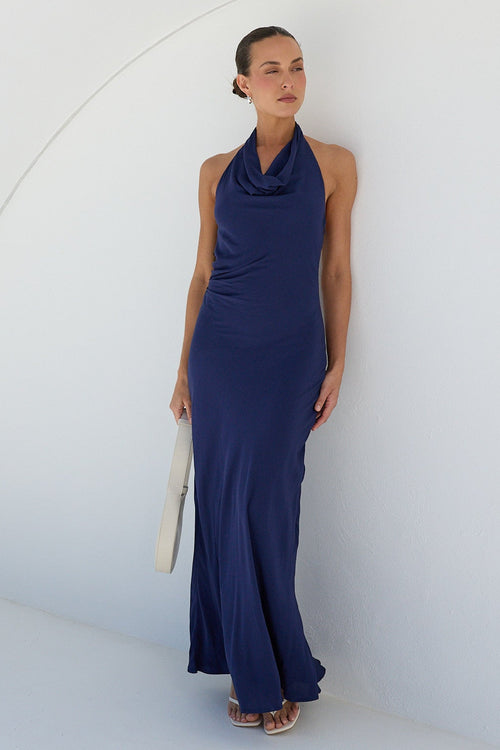 A woman wearing a sleeveless navy blue cowl-neck maxi dress stands inside a white arched alcove on a sunny day, holding a white clutch.