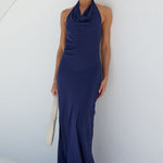 A woman wearing a sleeveless navy blue cowl-neck maxi dress, holding a white clutch bag, posing against a minimalist white wall with a curved architectural line.