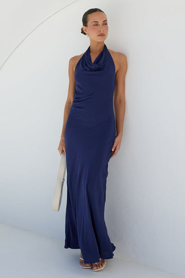 A woman wearing a sleeveless navy blue cowl-neck maxi dress, holding a white clutch bag, posing against a minimalist white wall with a curved architectural line.