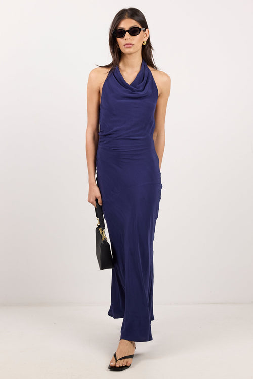 A woman modeling a sleeveless navy blue cowl-neck maxi dress with a draped front, paired with black sunglasses, a small black handbag, and black sandals, set against a plain white studio background.