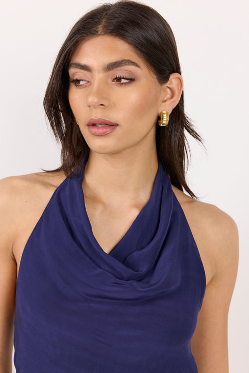 A woman wearing a navy blue halter-neck top with a draped cowl neckline, paired with gold hoop earrings, against a plain white background.