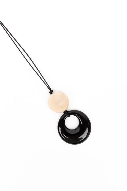 A minimalist pendant necklace featuring a glossy black circular donut pendant and a smooth, matte cream-colored round bead on a black cord, set against a plain white background.