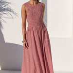 A woman modeling a sleeveless midi dress with a textured, patterned dusty rose bodice and a matching vertically striped skirt, set against a bright white stucco wall with an archway and a large terracotta planter.