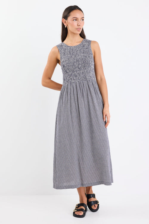 Woman wearing a navy mini gingham midi dress with a shirred bodice, sleeveless crew neckline, and relaxed gathered skirt.