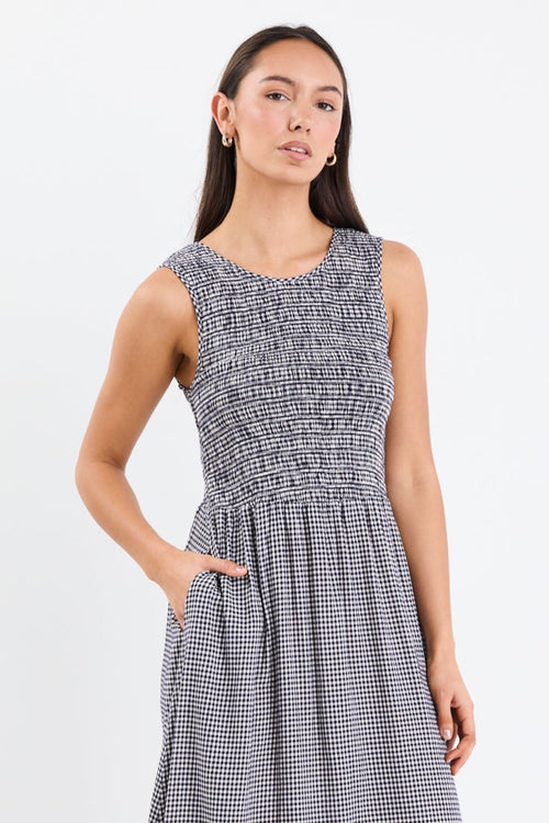 Woman wearing a navy mini gingham midi dress with a shirred bodice, sleeveless crew neckline, and relaxed gathered skirt.