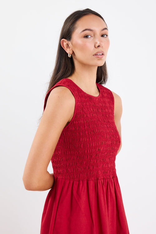 Woman wearing a sleeveless plum midi dress with a shirred bodice, crew neckline, and gathered flowing skirt.