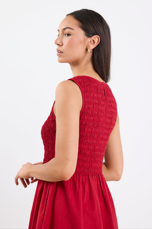 Woman wearing a sleeveless plum midi dress with a shirred bodice, crew neckline, and gathered flowing skirt.