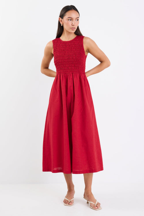 Woman wearing a sleeveless plum midi dress with a shirred bodice, crew neckline, and gathered flowing skirt.