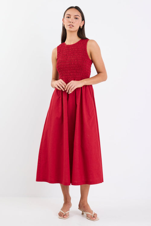 Woman wearing a sleeveless plum midi dress with a shirred bodice, crew neckline, and gathered flowing skirt.