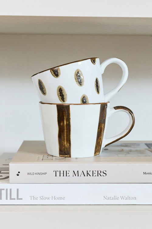 Two stacked white ceramic mugs with black and gold speckled patterns, displayed on a stack of books on a white shelf.