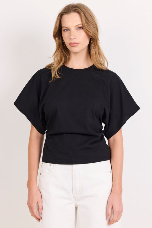 A woman modeling a black short-sleeve crop top with a gathered waist and white pants, standing against a plain off-white studio background.