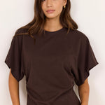 A woman wearing a dark brown short-sleeve t-shirt with draped, balloon sleeves and beige pants, standing against a plain off-white background.