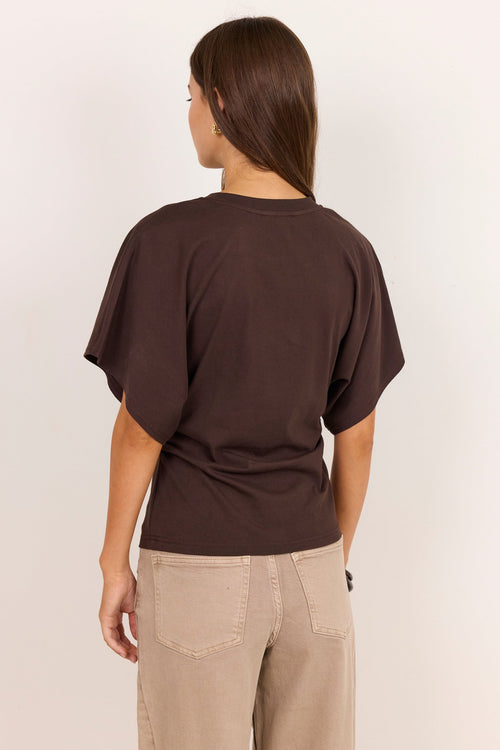 A woman viewed from the back wearing a dark brown short-sleeve t-shirt and beige pants, standing against a plain off-white background.