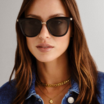 model wears cat-eye sunglasses featuring a sleek black frame paired with khaki-tinted lenses. A slim gold-tone bridge and matching gold arm accents elevate the look with a modern, minimalist touch.