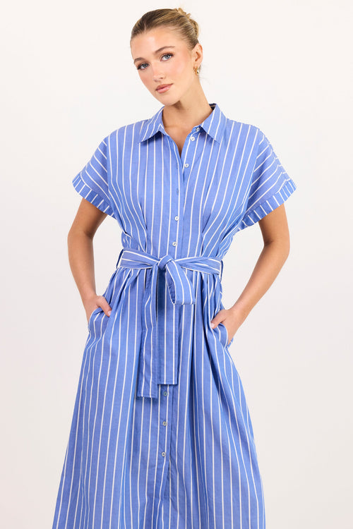 A woman modeling a blue and white vertically striped shirt dress with a tie waist belt, standing against a plain off-white studio background.