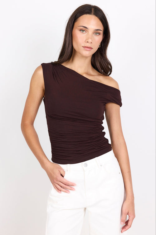 A woman modeling a dark brown off-the-shoulder ruched top paired with white pants, photographed against a clean white studio background.