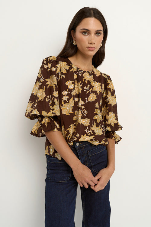 Woman wearing a chocolate brown floral blouse with short puff sleeves and dark denim jeans.