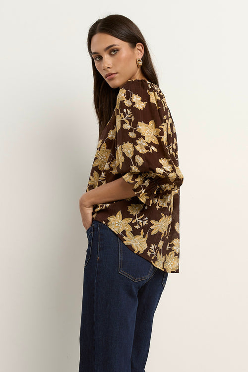 Woman wearing a chocolate brown floral blouse with short puff sleeves and dark denim jeans.