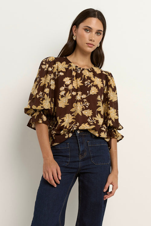 Woman wearing a chocolate brown floral blouse with short puff sleeves and dark denim jeans.