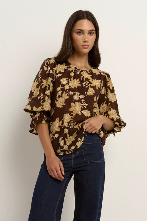 Woman wearing a chocolate brown floral blouse with short puff sleeves and dark denim jeans.
