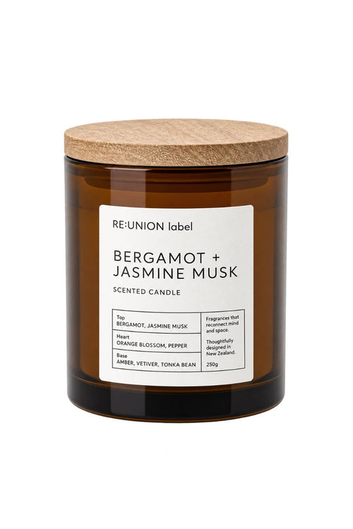 A Re:Union candle in an amber glass jar with a light wood lid, featuring a white label that reads 'BERGAMOT + JASMINE MUSK'.