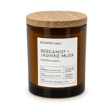A 135g scented candle in an amber glass jar with a light wood lid, featuring a white label from the brand RE:UNION label. The scent is Bergamot + Jasmine Musk.