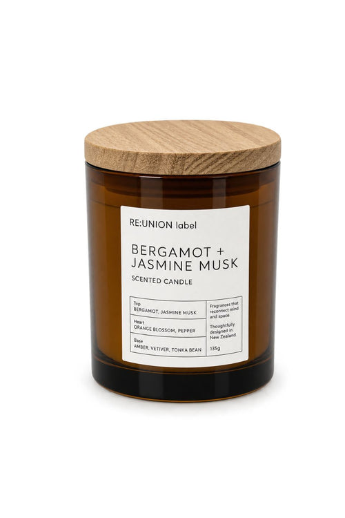 A 135g scented candle in an amber glass jar with a light wood lid, featuring a white label from the brand RE:UNION label. The scent is Bergamot + Jasmine Musk.