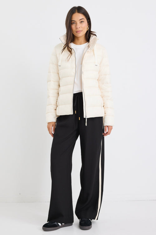 Woman wearing an ivory zip-up puffer jacket with a high neck, paired with black track pants and a white tee, leaning casually against a white wall.