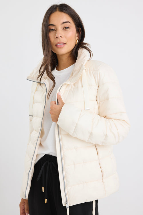 Woman wearing an ivory zip-up puffer jacket with a high neck, paired with black track pants and a white tee, leaning casually against a white wall.