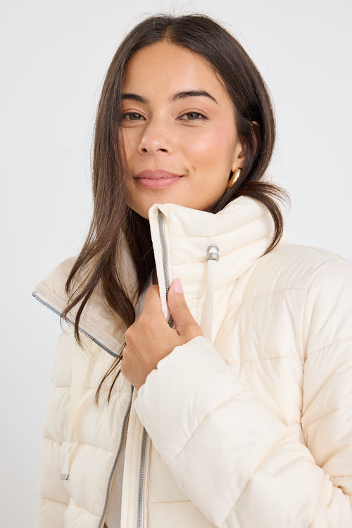 Woman wearing an ivory zip-up puffer jacket with a high neck, paired with black track pants and a white tee, leaning casually against a white wall.