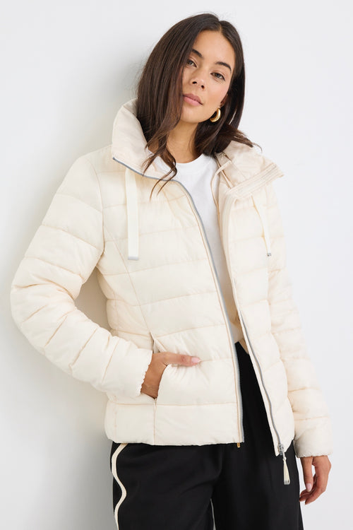 Woman wearing an ivory zip-up puffer jacket with a high neck, paired with black track pants and a white tee, leaning casually against a white wall.