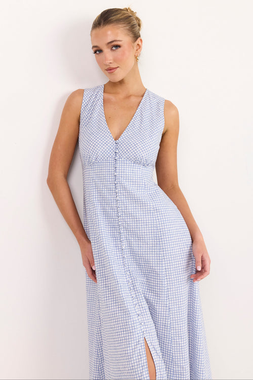 A woman modeling a sleeveless light blue and white gingham maxi dress with a V-neckline and front button detail, standing against a plain white wall.