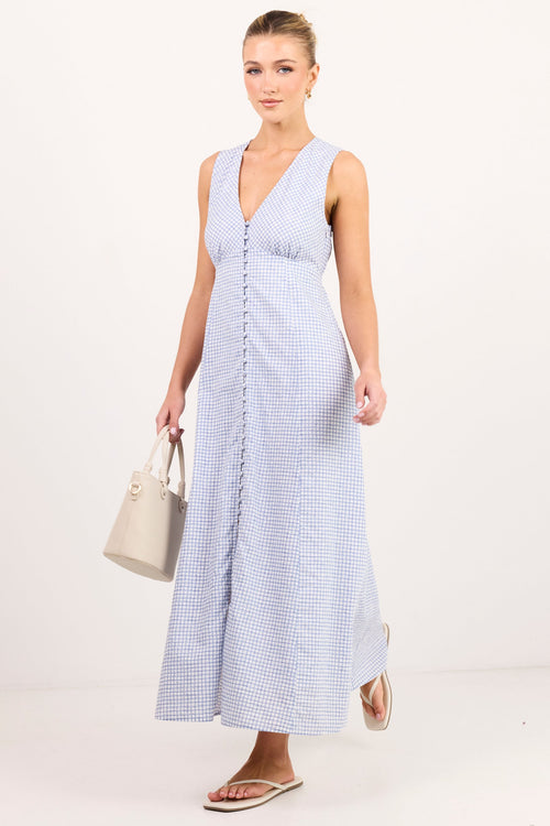 A woman modeling a sleeveless light blue and white gingham maxi dress with a V-neckline and front button detail, holding a beige handbag against a plain white studio background.